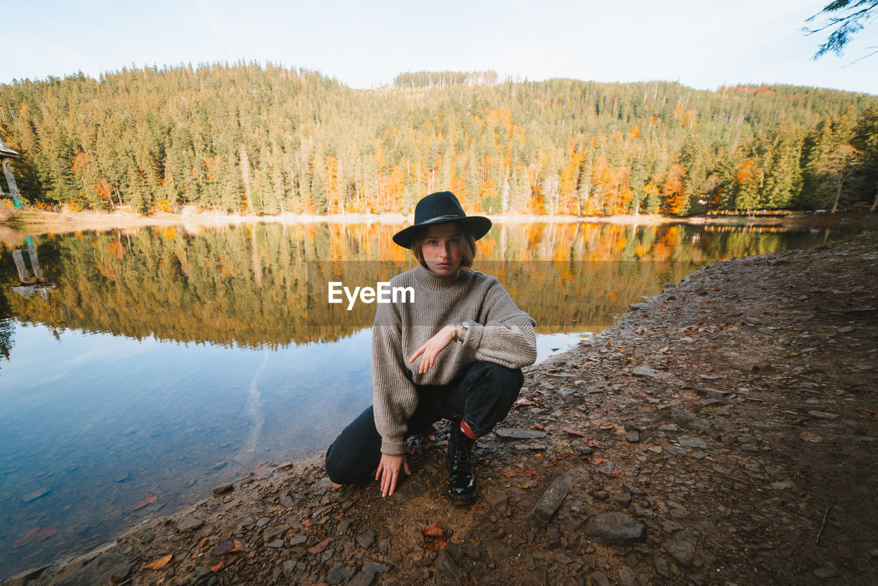 Young cool female tourist in trendy apparel looking at camera from dry coast against transparent water reflecting autumn trees