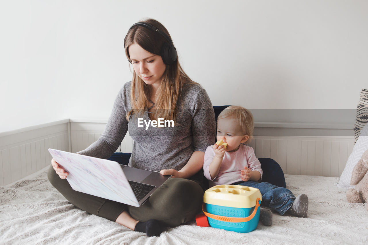 Mother using laptop while sitting with baby girl on bed at home