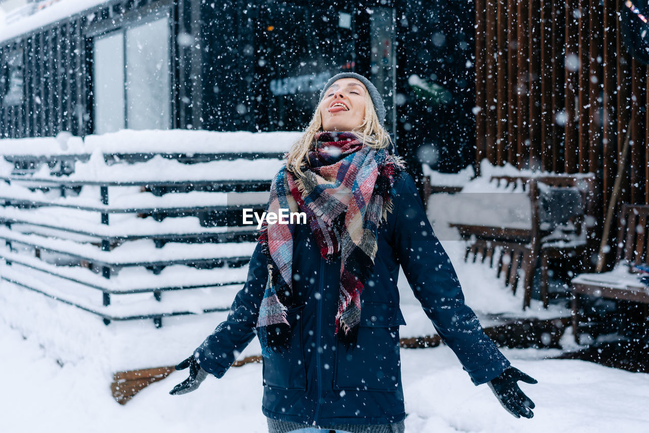 A blonde woman standing under a snowfall catches snowflakes with her tongue like a child having fun.