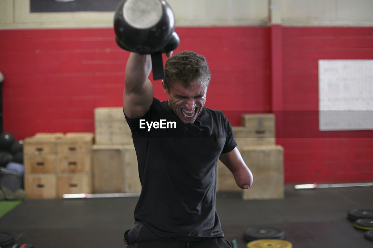 Aggressive male adaptive athlete lifting kettlebell while standing against wall in gym