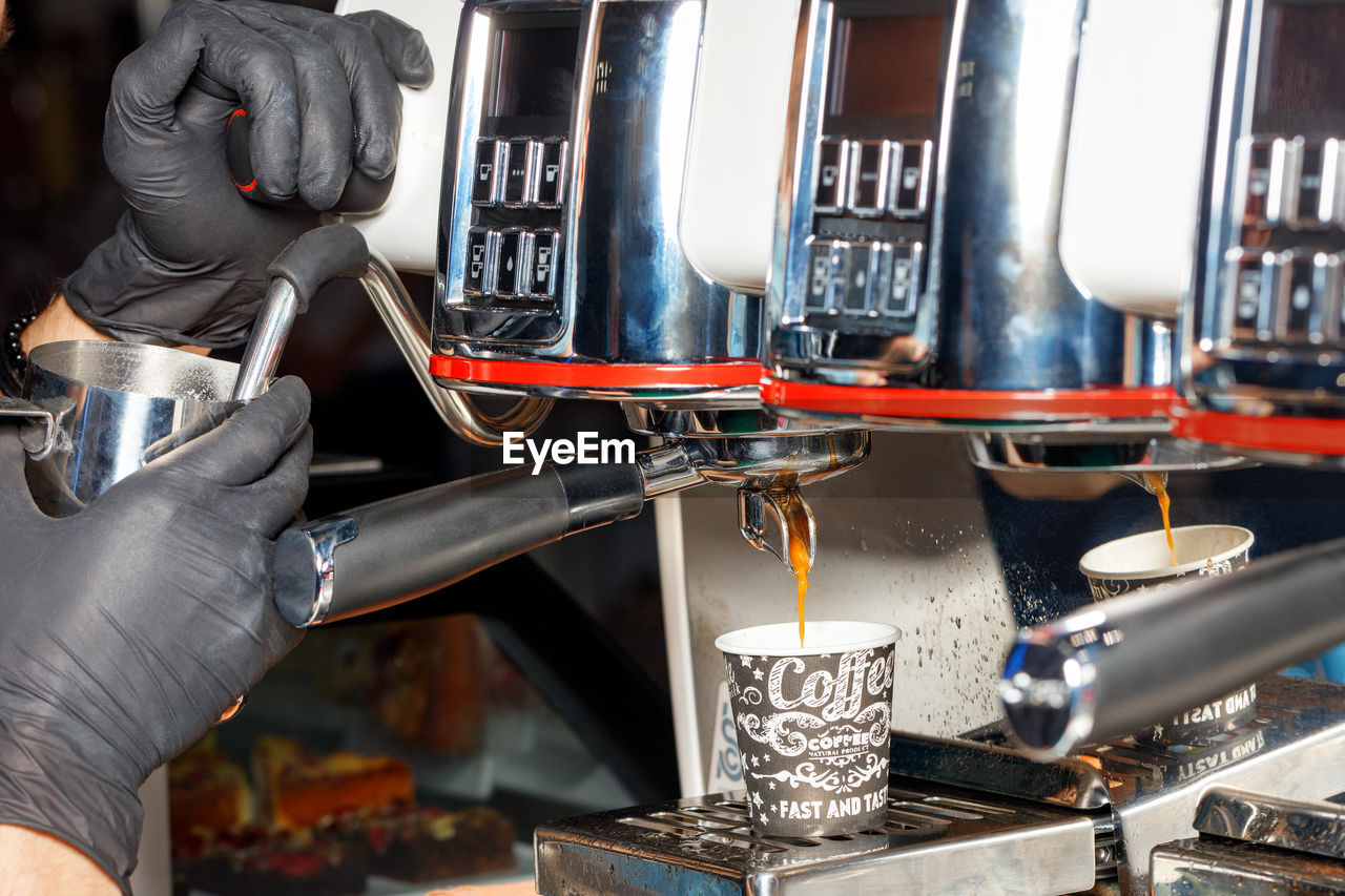 A barista in black rubber gloves pours coffee into a paper cup from a coffee machine.