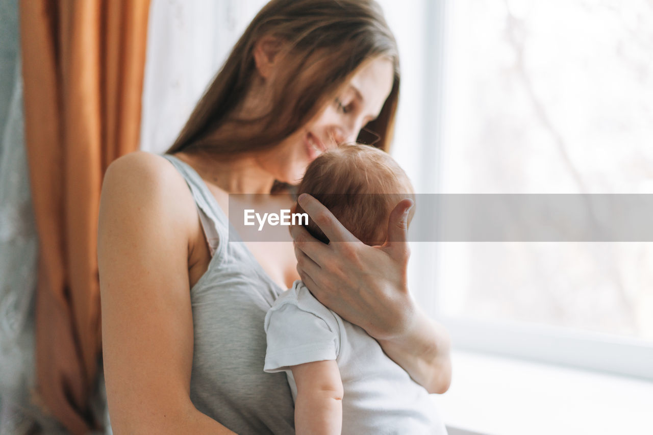 Young woman mom with long hair holding baby girl on hands near window at home