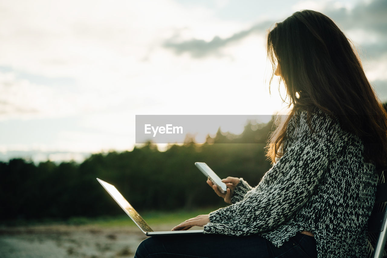 Mid adult woman with laptop text messaging while sitting at beach during sunset