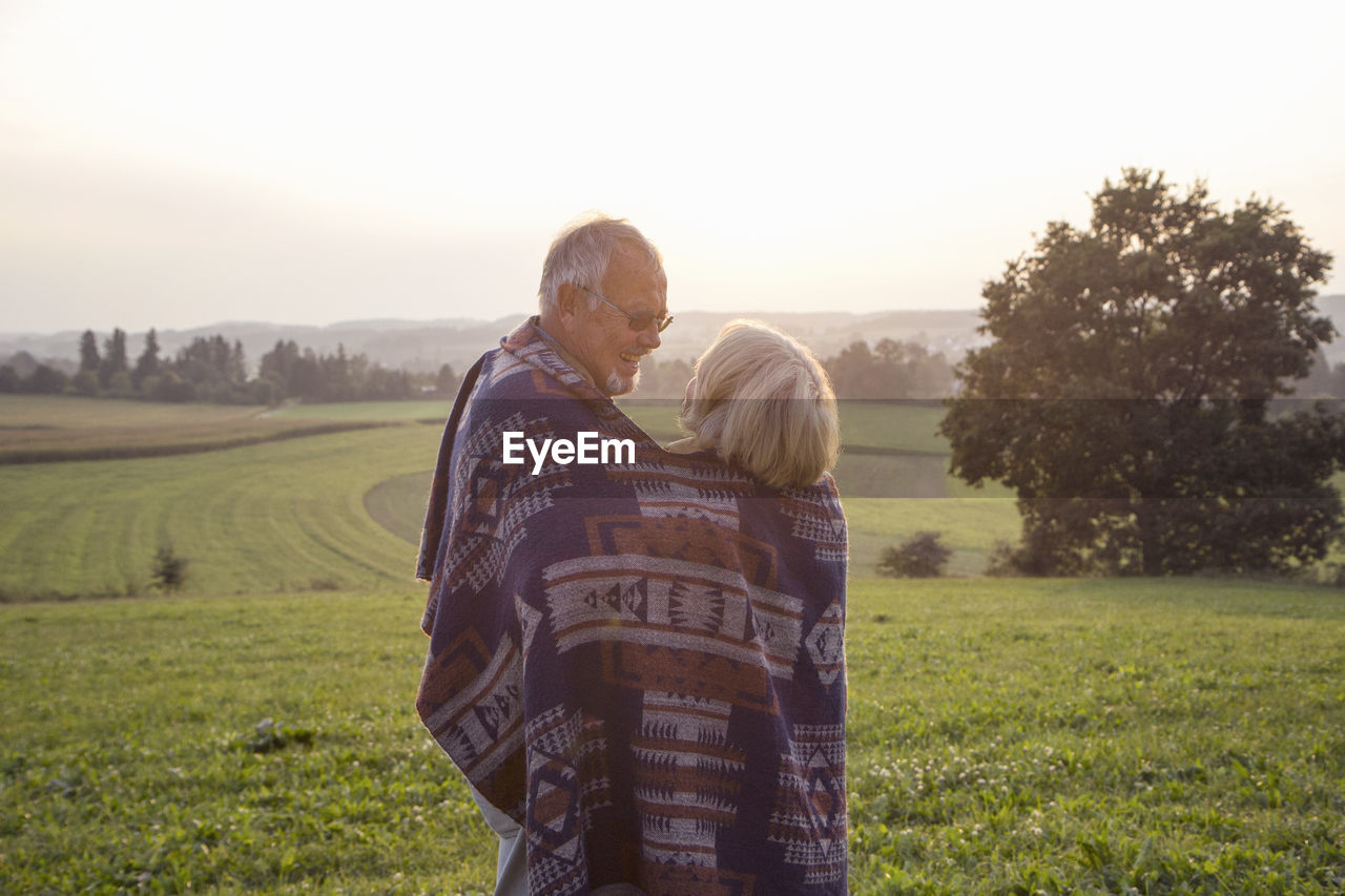 Happy senior couple standing on a meadow at sunset sharing blanket