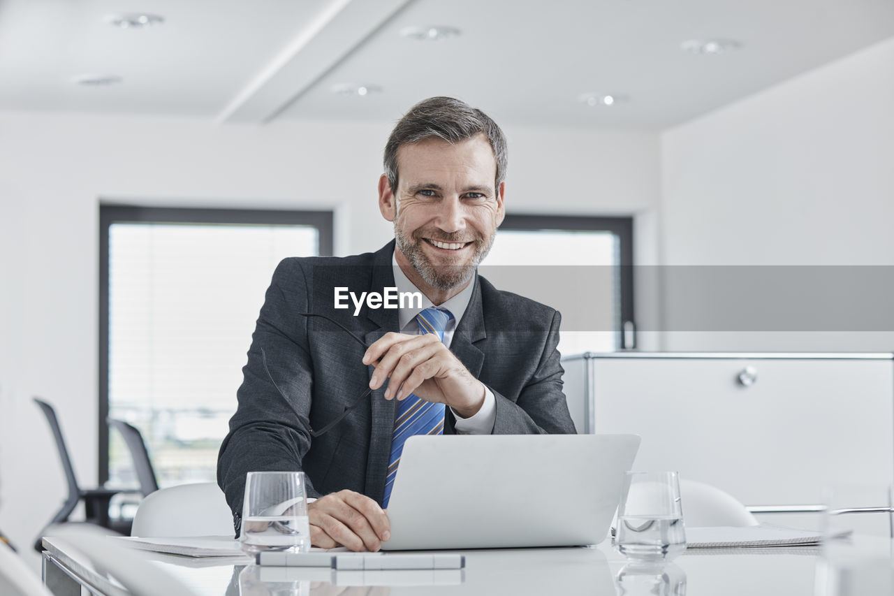 Portrait of smiling businessman with laptop at desk in office