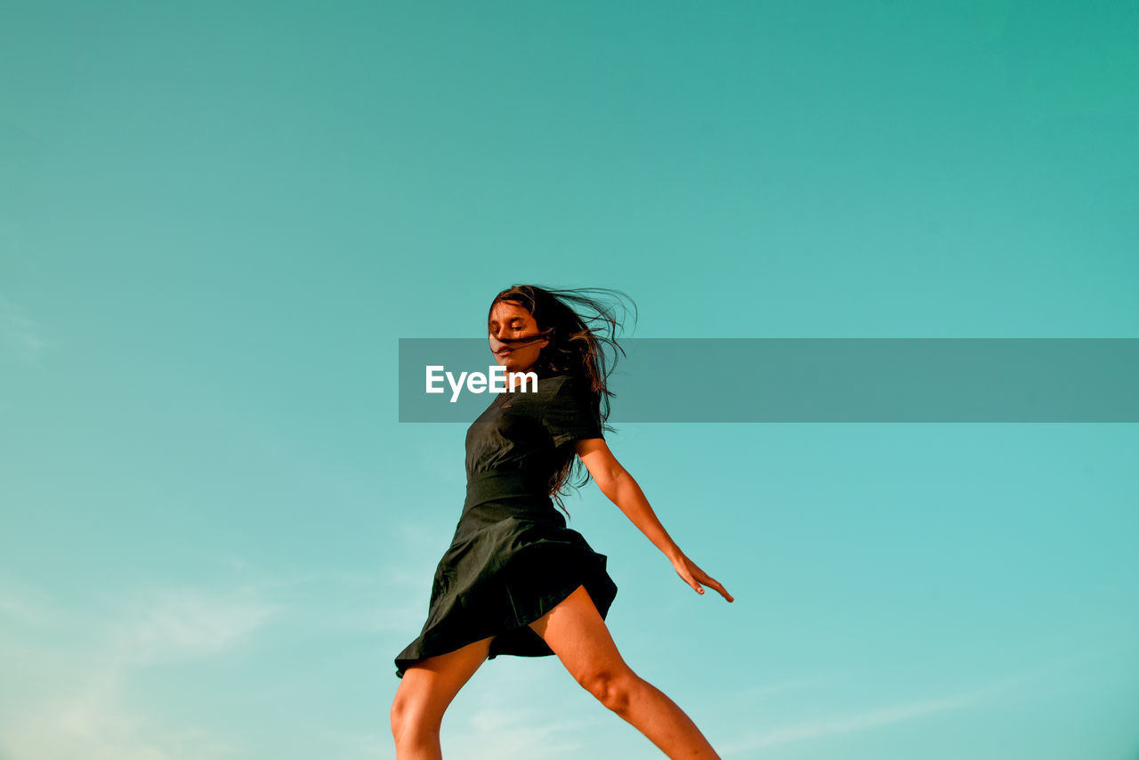Low angle view of woman jumping against clear blue sky