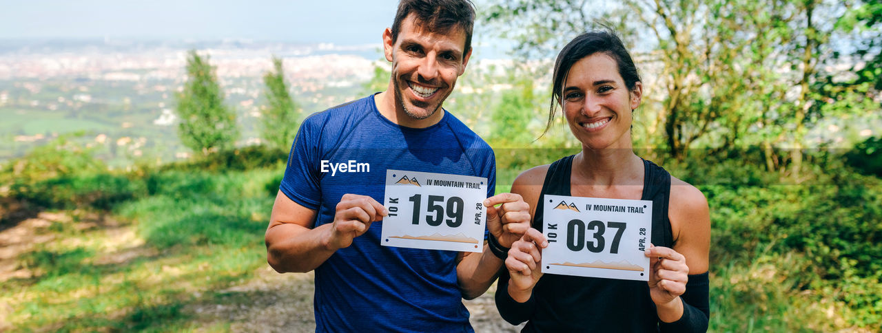Portrait of happy couple holding marathon bibs while standing on field