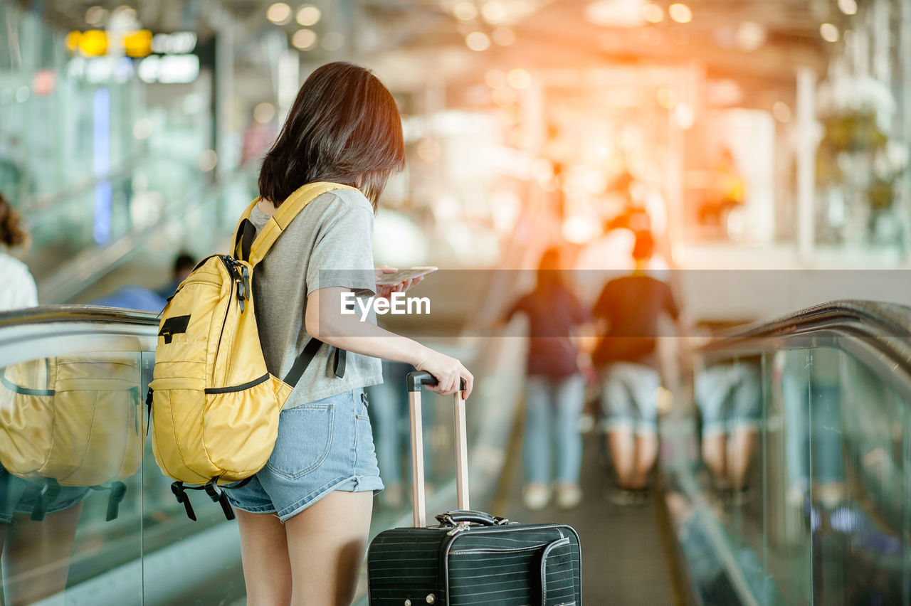 Rear view of woman with luggage using smart phone while standing on moving walkway