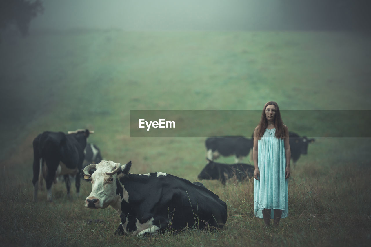 Portrait of woman standing with cows on field during foggy weather