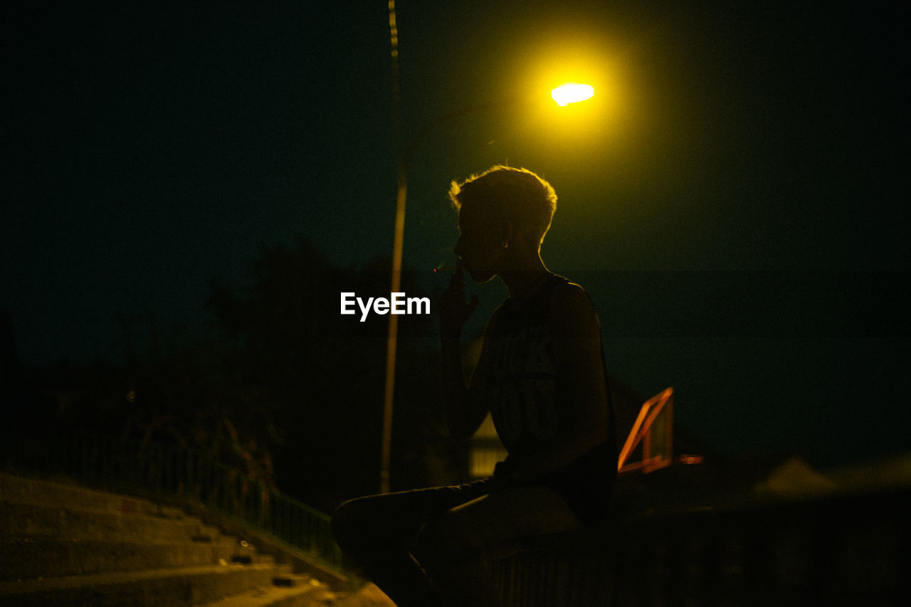 Silhouette woman smoking cigarette while sitting on railing against illuminated street light at night