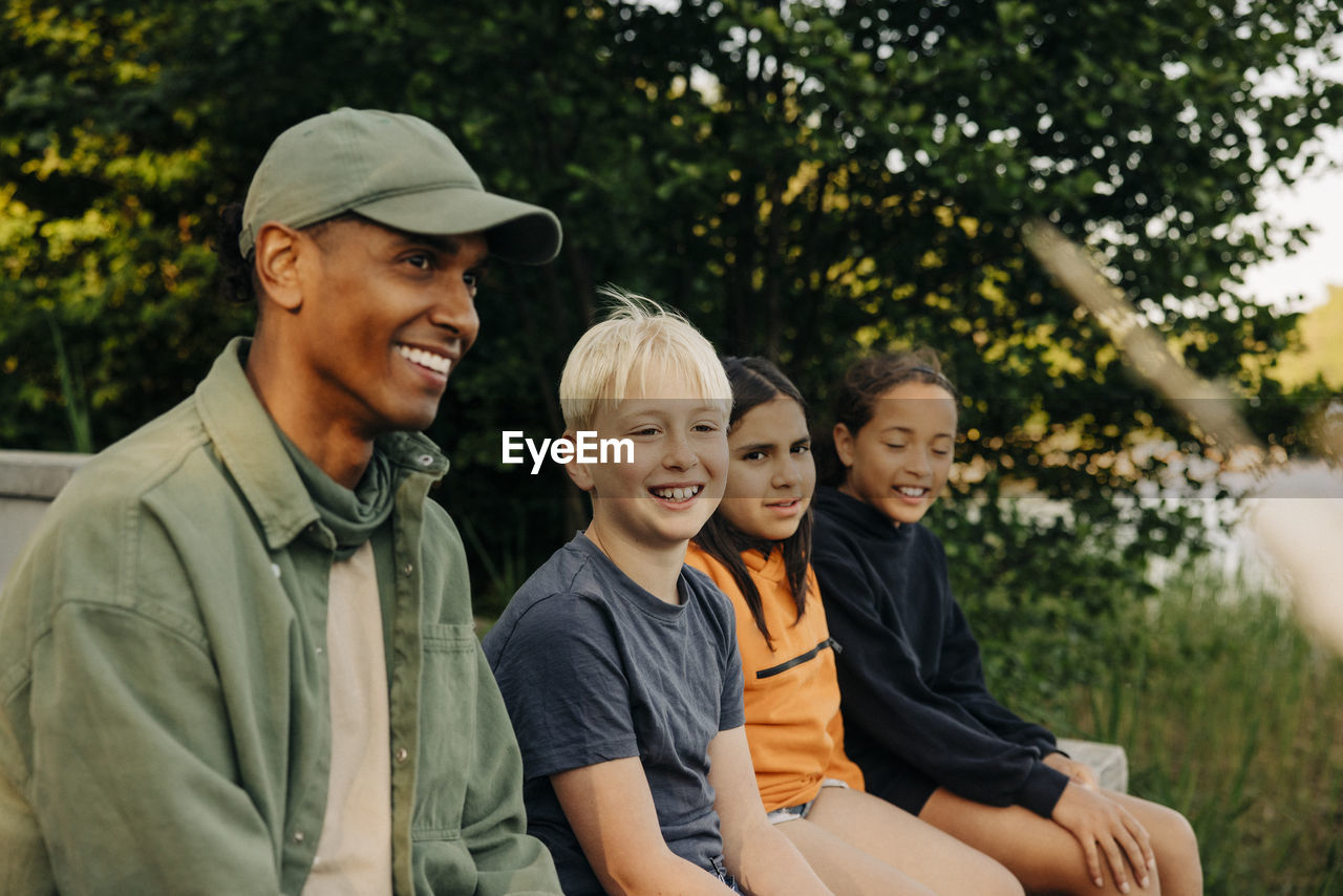 Kids sitting with smiling counselor wearing cap at summer camp