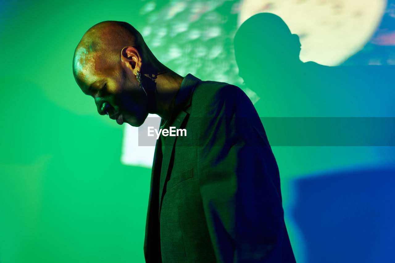 Side view of confident bald african american male model with necklace and earring in trendy outfit standing with eyes closed at wall with glowing lights in modern studio