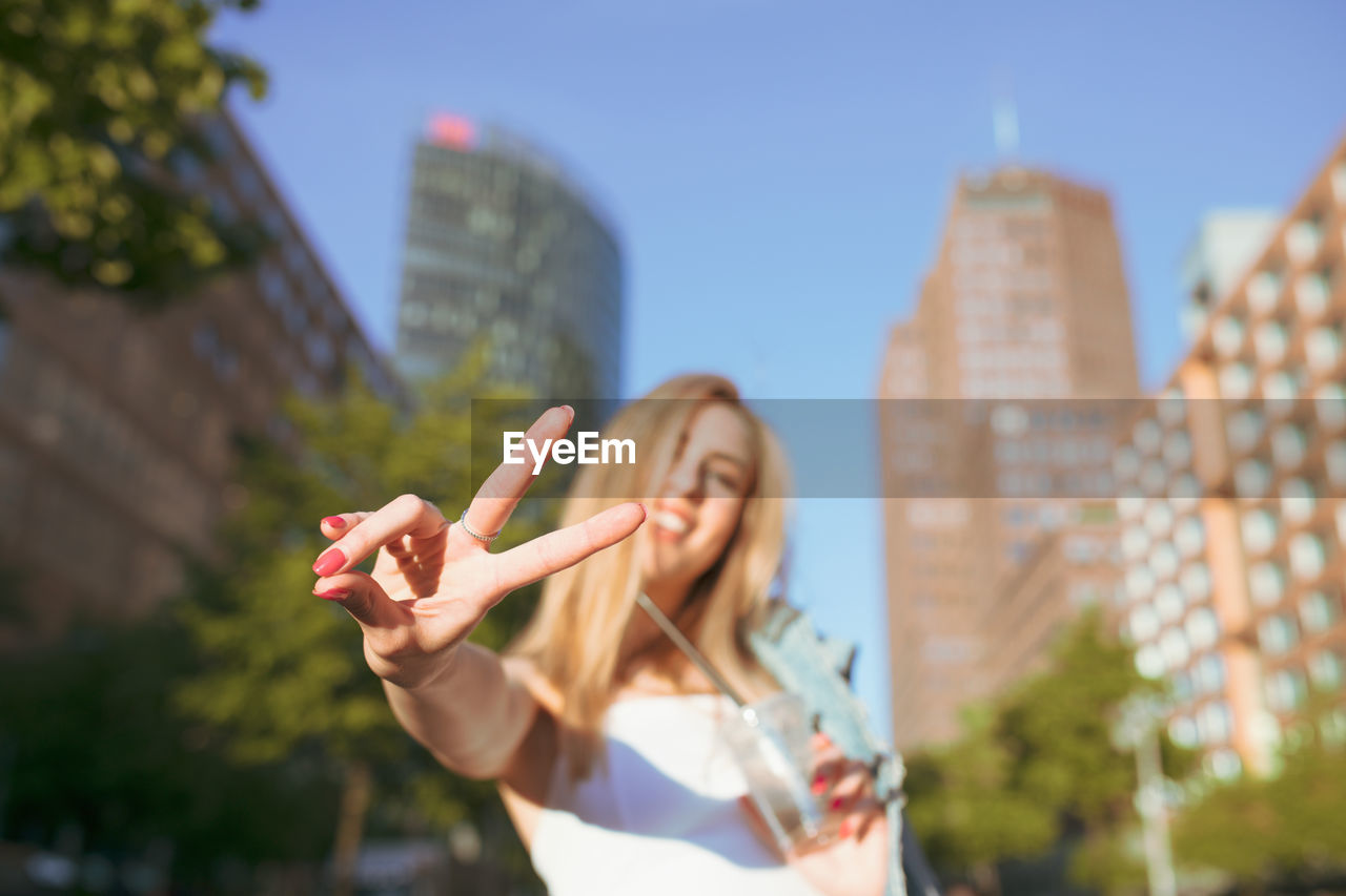 Portrait of smiling woman showing peace sign against buildings in city