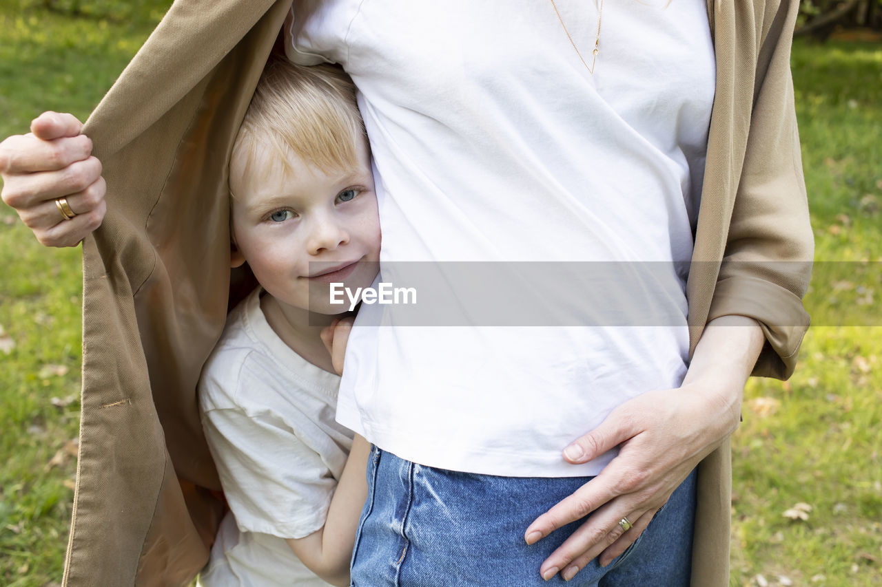 Little smiling caucasian 7 yo boy looks out mother's jacket, hiding behind her back in park. mom's