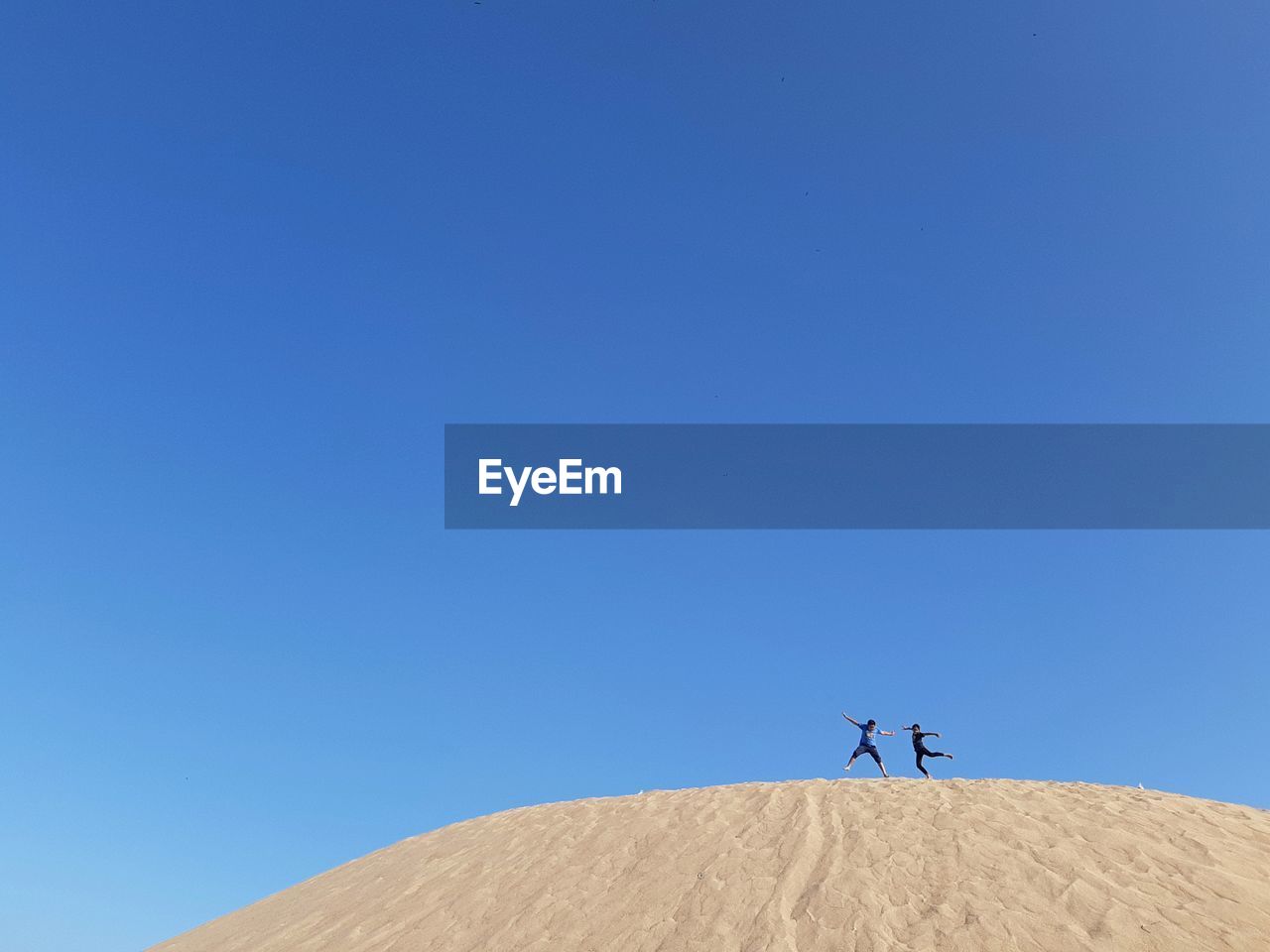 Low angle view of boys jumping on sand dune at desert against clear blue sky