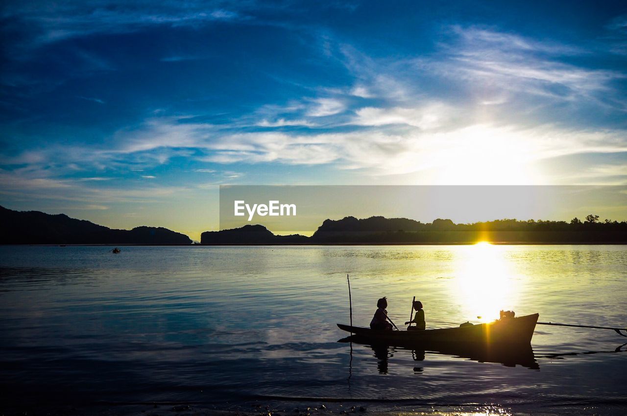 Silhouette people sitting on rowboat in lake against sky during sunset