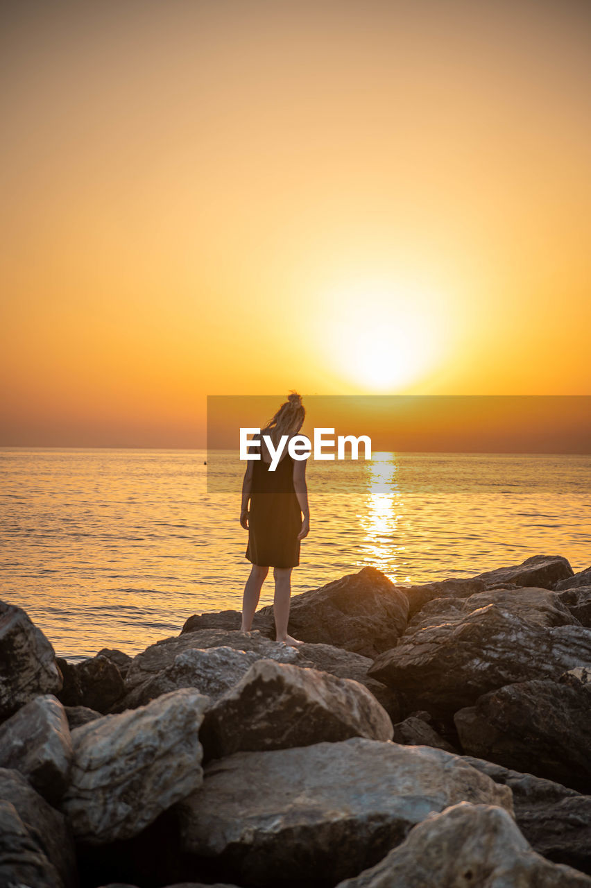 REAR VIEW OF MAN STANDING ON ROCK AGAINST SEA