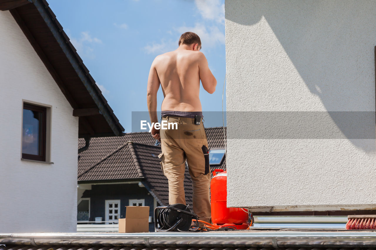 REAR VIEW OF SHIRTLESS MAN STANDING BY BUILDING AGAINST SKY
