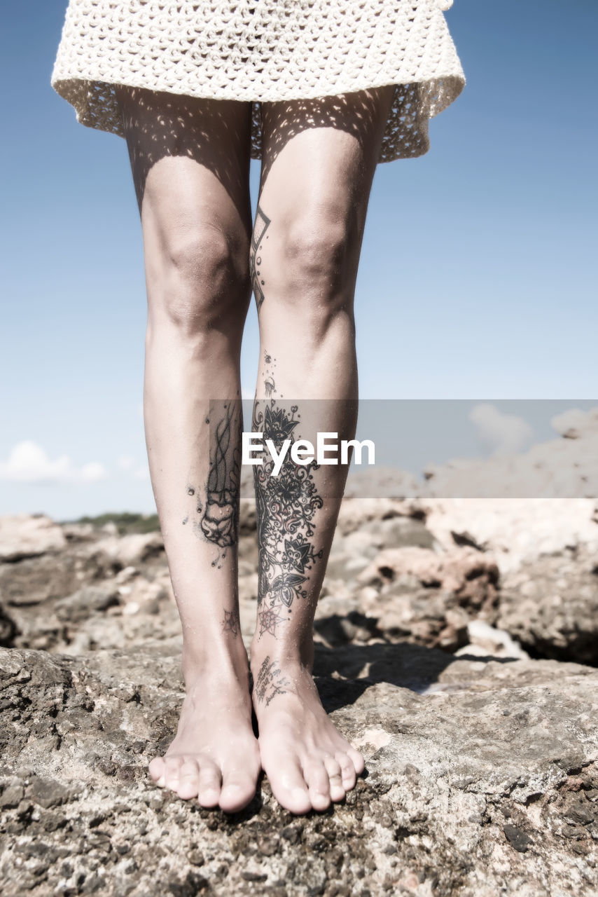 Low section of woman with tattooed legs standing on rock against sky