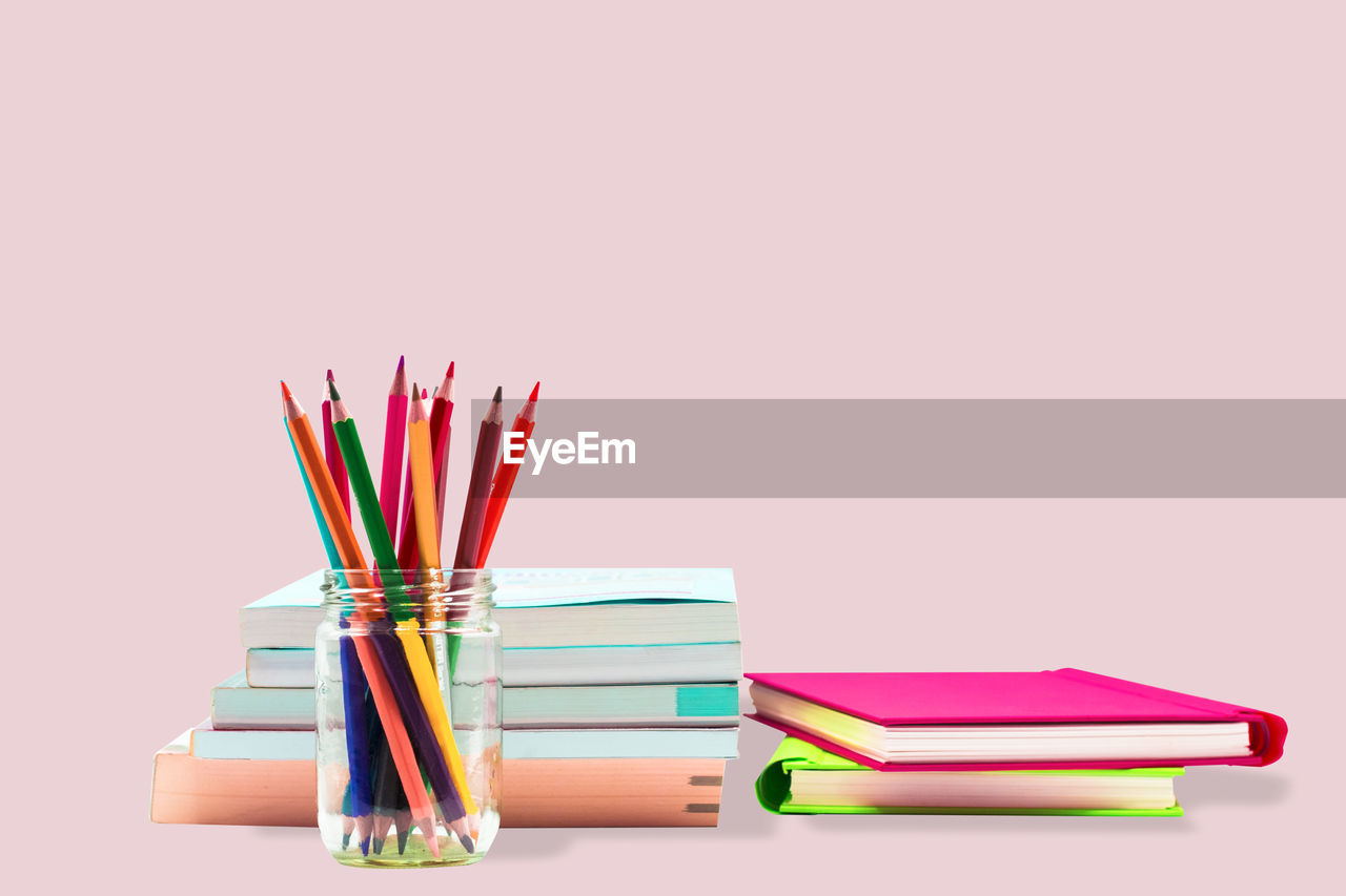 Close-up of multi colored pencils in jar with book against pink background