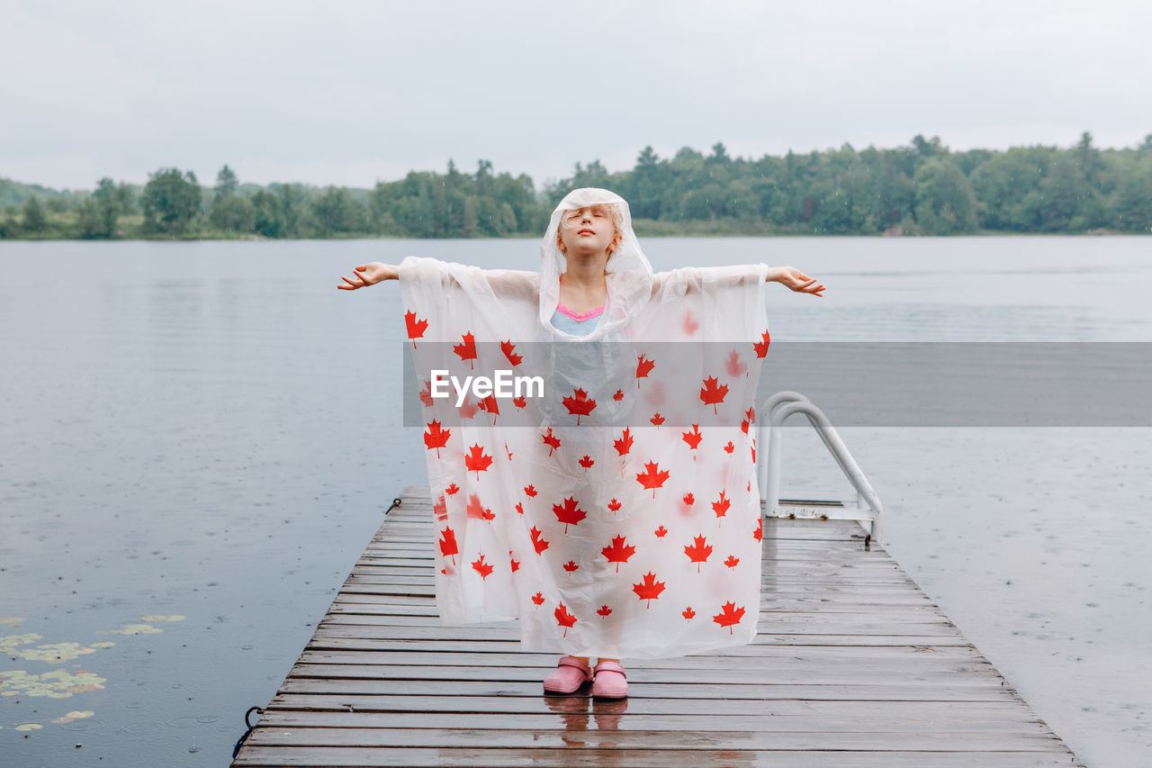Girl wearing raincoat standing on pier with arms outstretched
