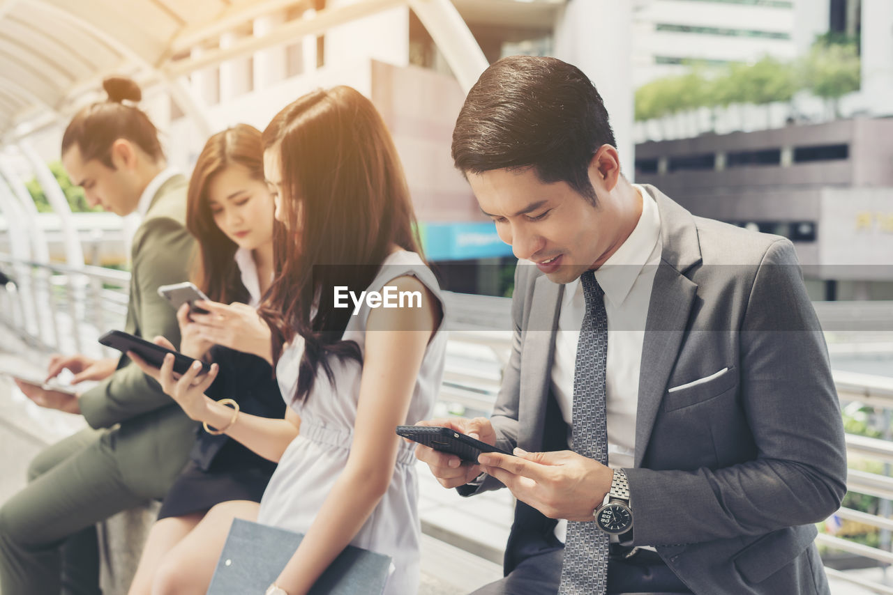 Colleagues using phones while sitting against buildings in city