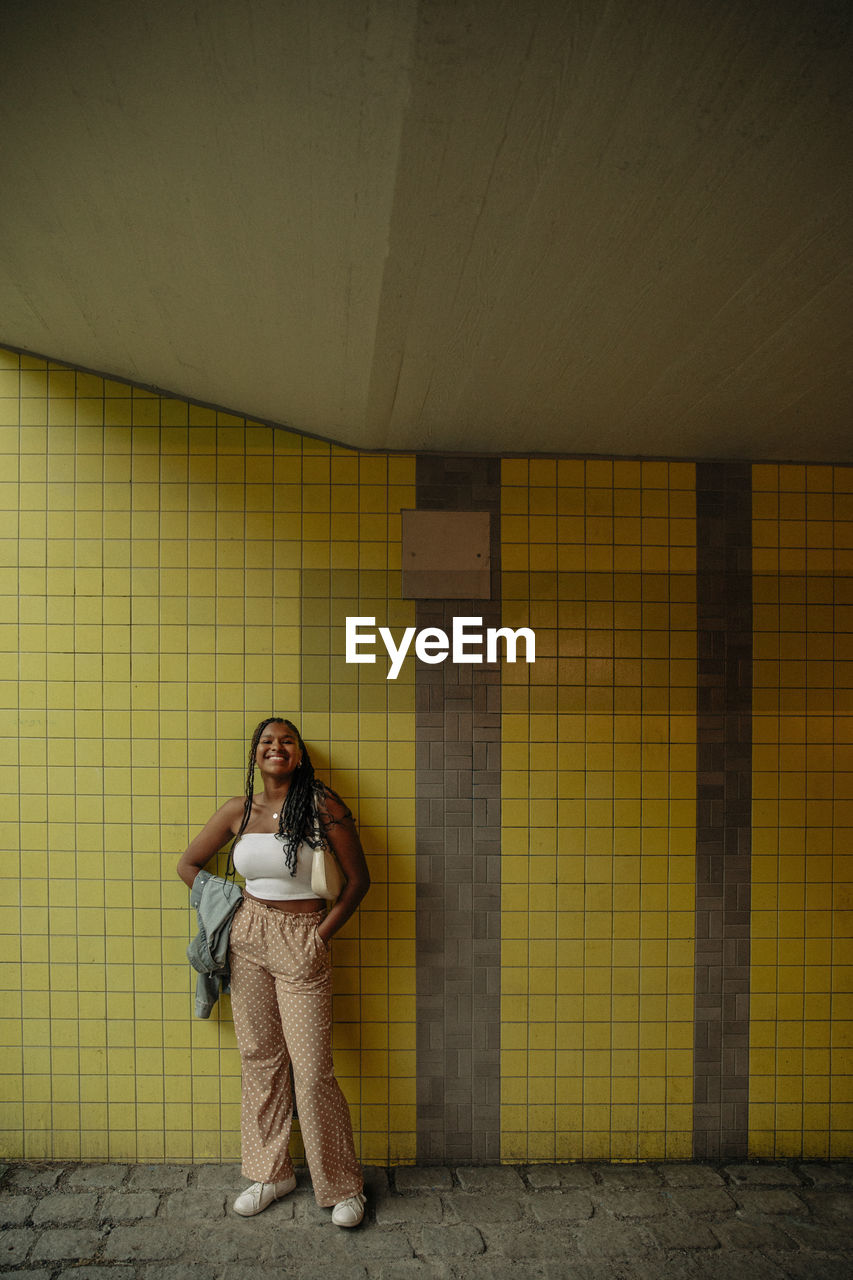 Full length of smiling teenage girl standing with hands in pockets against yellow wall