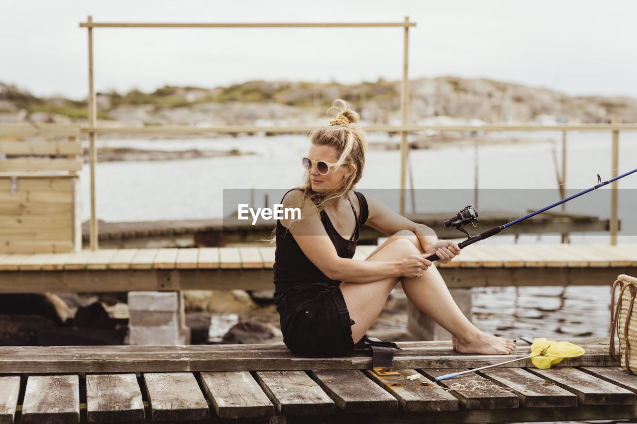 Mature woman with fishing rod looking over shoulder while sitting on jetty at harbor