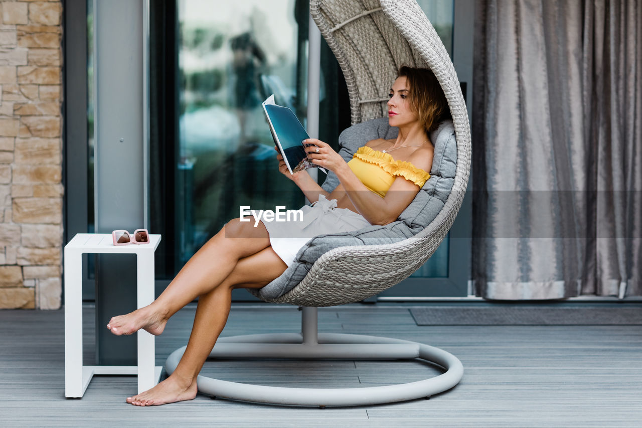 A young woman is sitting in a hanging chair on the terrace and reading a magazine