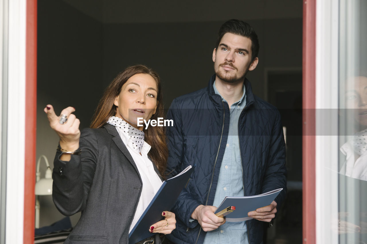 Female realtor and young man standing by window while pointing away