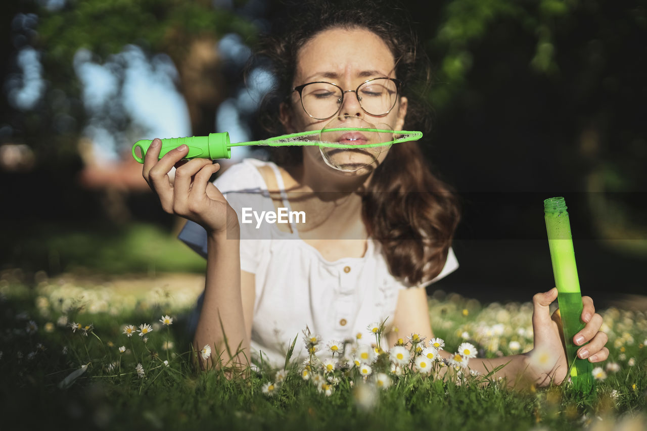 Portrait of a beautiful caucasian teenager girl blowing soap bubbles while lying on a flower meadow