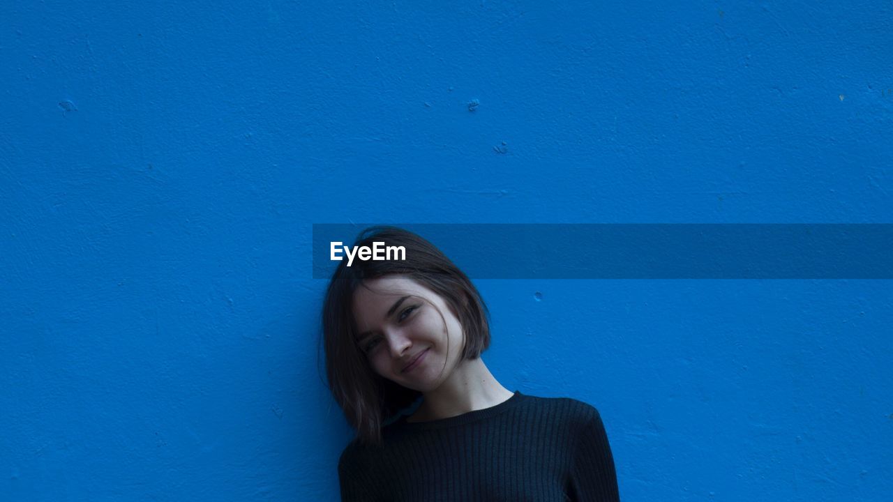 Portrait of young woman smiling while standing by blue wall