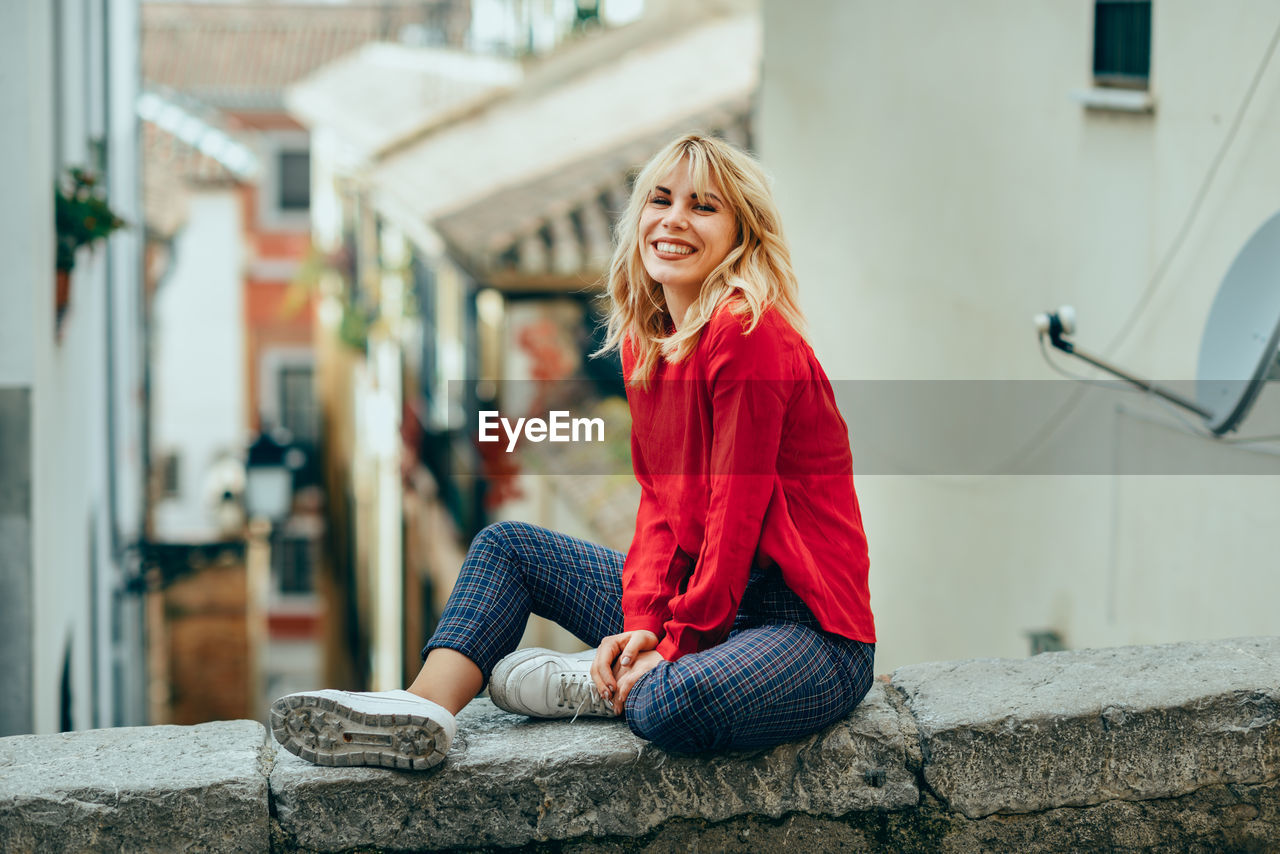 Portrait of cheerful young woman sitting on retaining wall in town