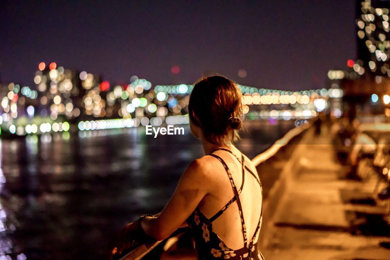 Rear view of woman leaning on promenade railing by river at night