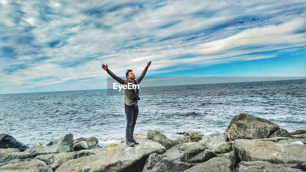 Man with arms raised standing on rocks at beach against sky