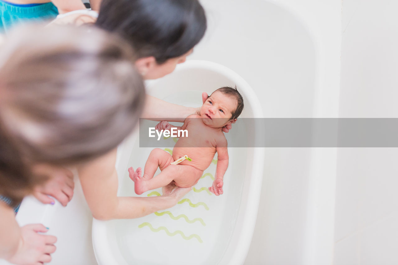 From above unrecognizable parent and kid washing crying newborn baby in warm water in basin at home