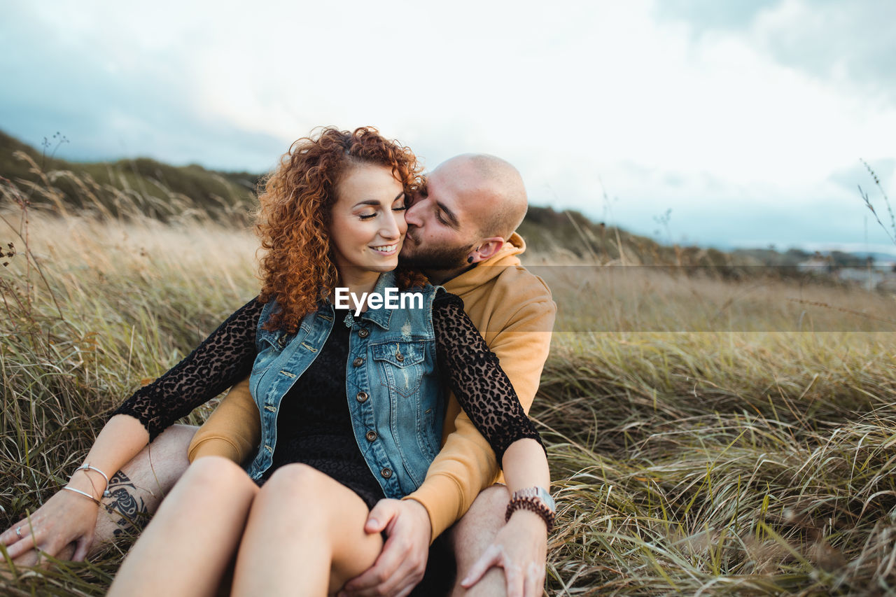 Young bald bearded man in yellow hoodie and jeans shorts embracing happy curly haired girlfriend in dress and denim vest while sitting together on meadow and talking