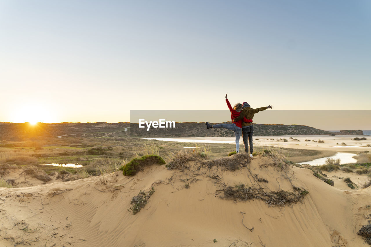 Young carefree couple with hand raised standing by man on sand dune during sunset