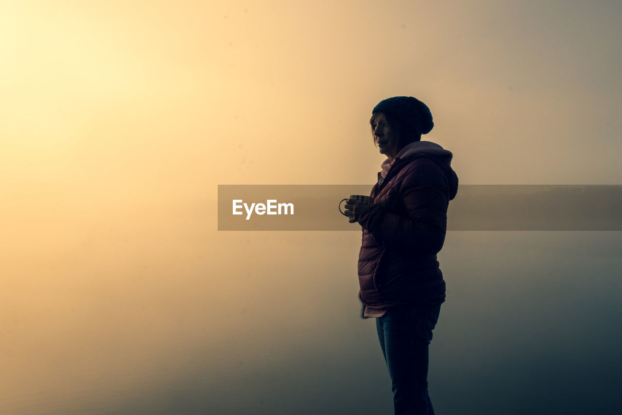 Side view of woman standing by lake against sky during dawn