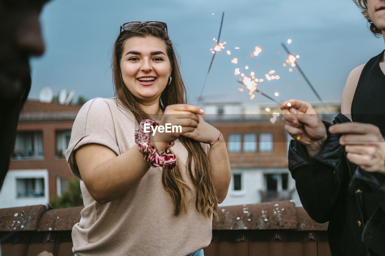 Portrait of smiling young woman holding sparklers with male friends on rooftop