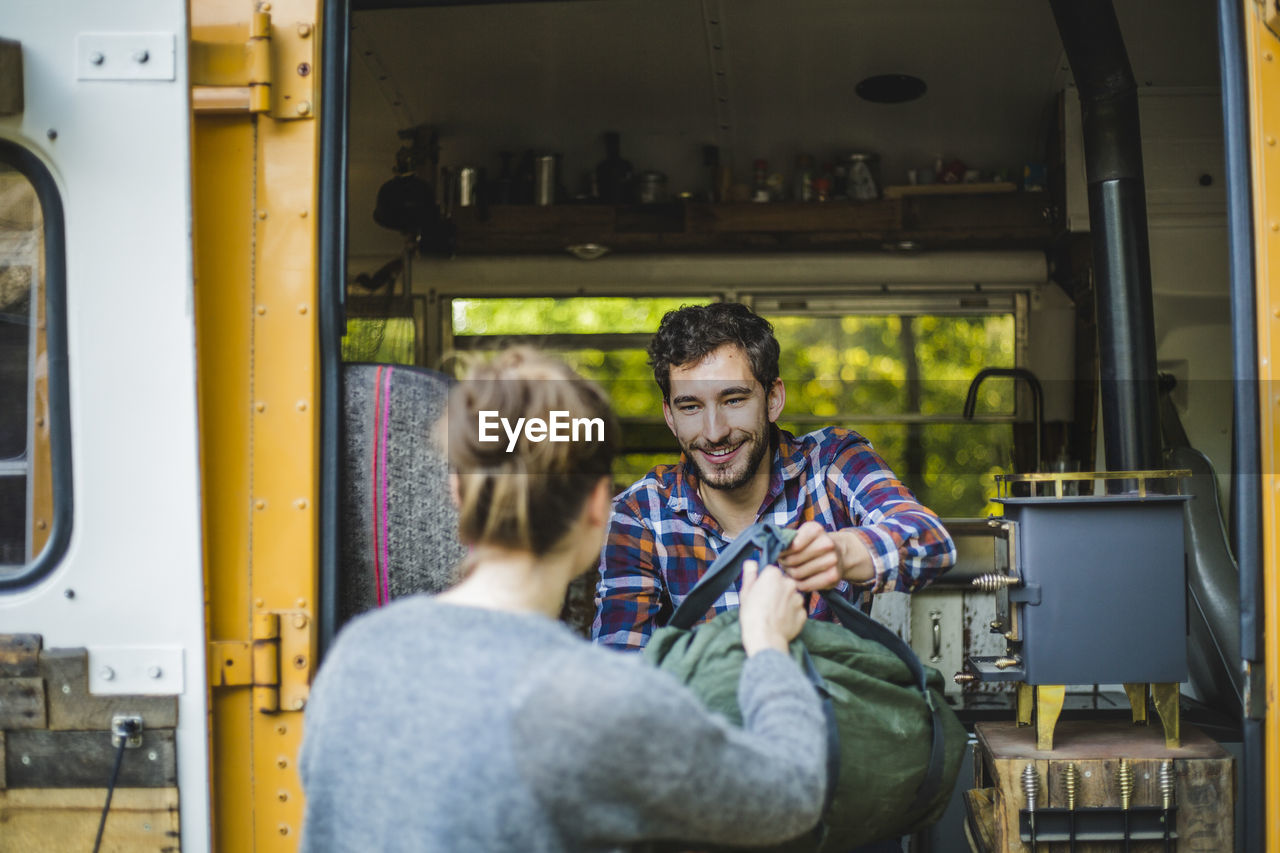 Smiling man giving luggage to woman for unloading from caravan during camping