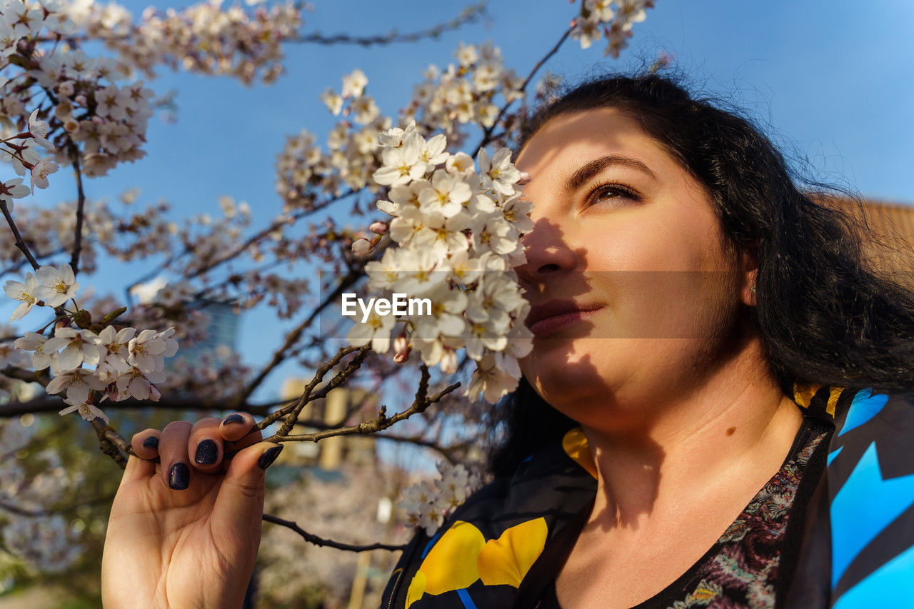Attractive plus size young woman sniffing cherry blossom branch in park in spring spring