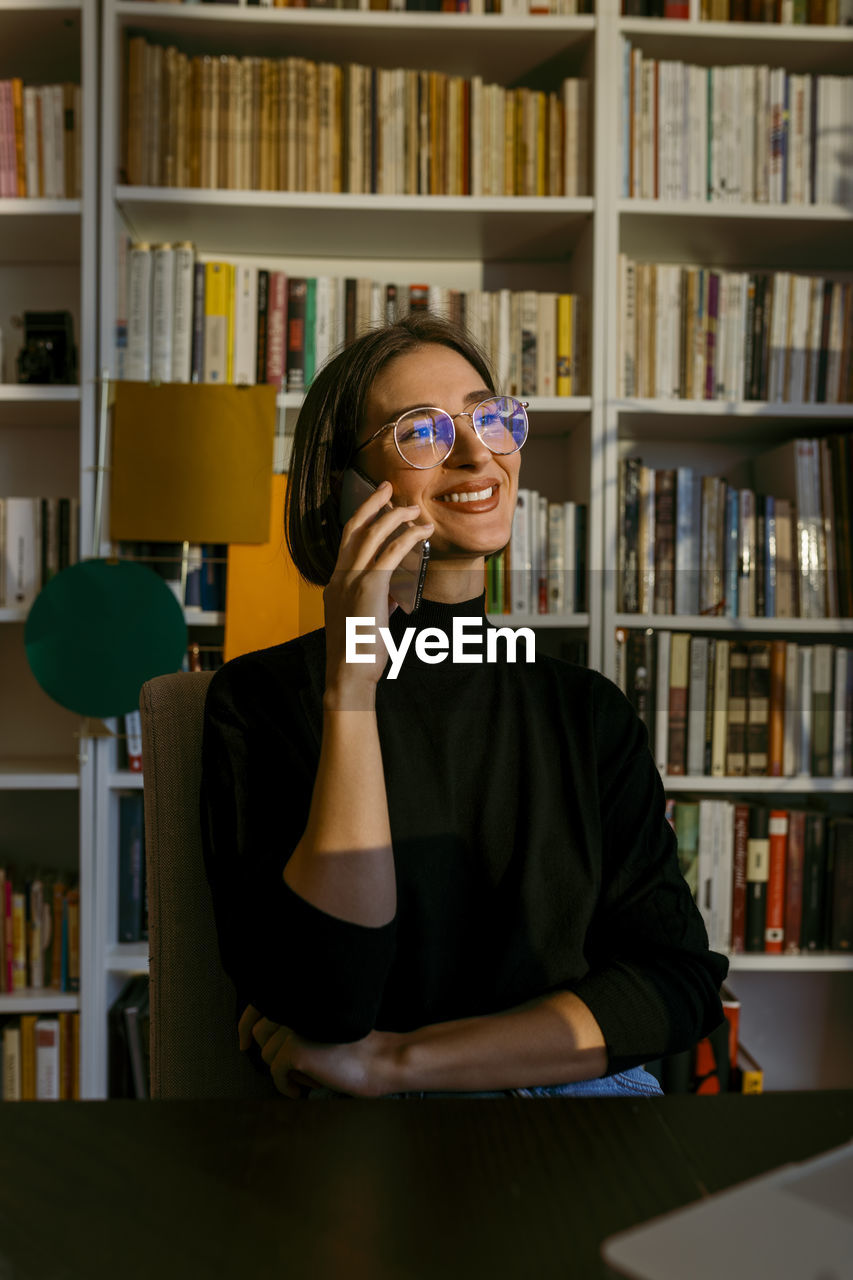 Smiling businesswoman looking away while talking on smart phone sitting at home