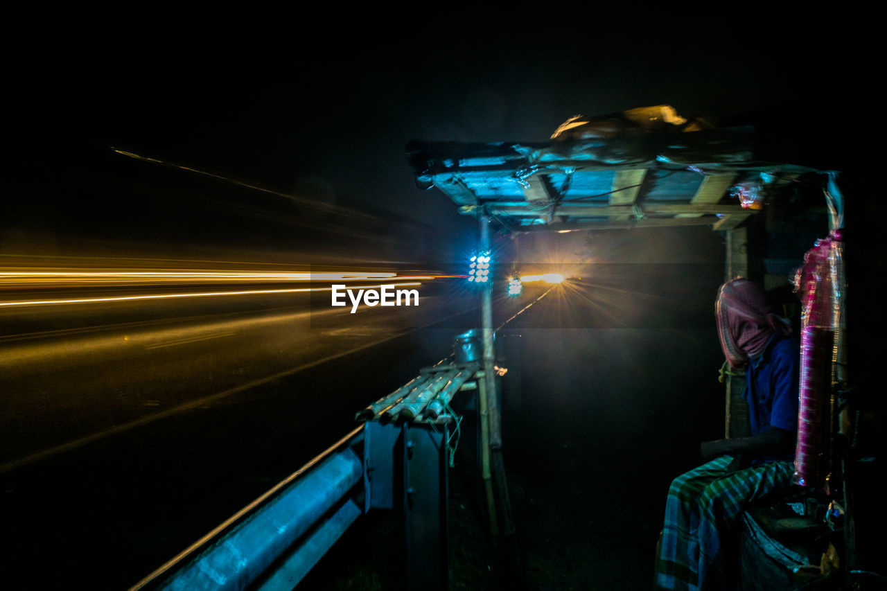 Man sitting at tea stall by light trails at night