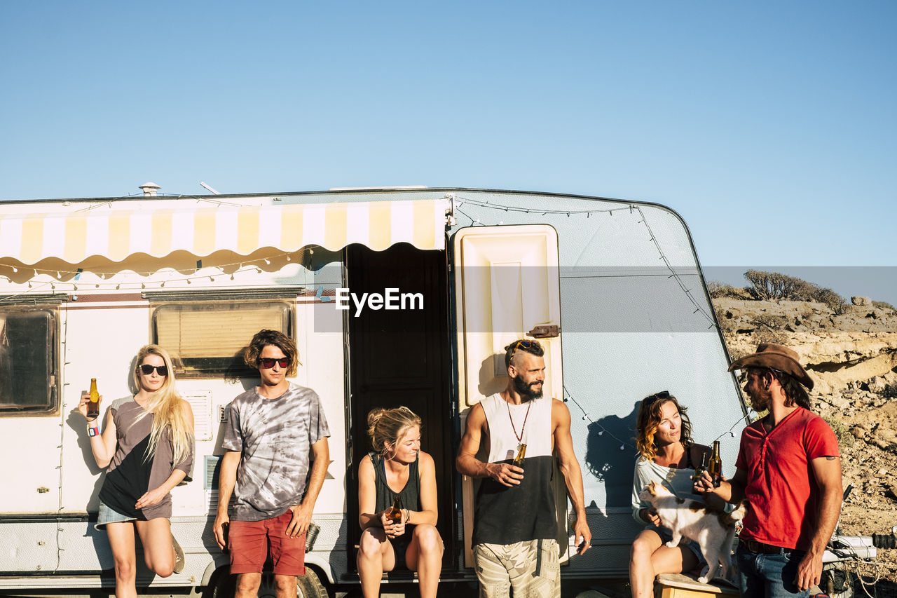 Friends holding beer bottles standing by motor home against sky