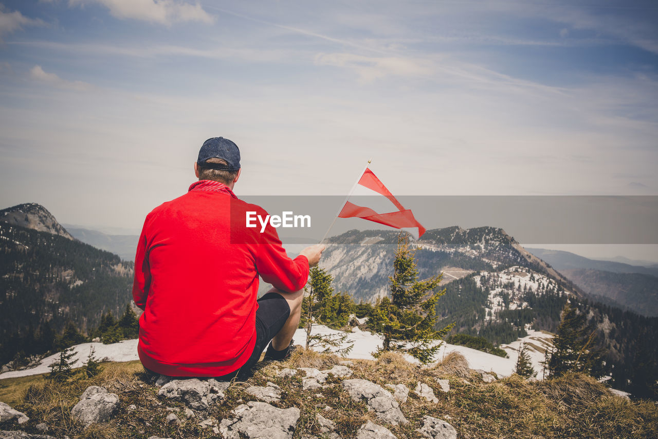 Rear view of man holding flag while sitting on mountain against sky