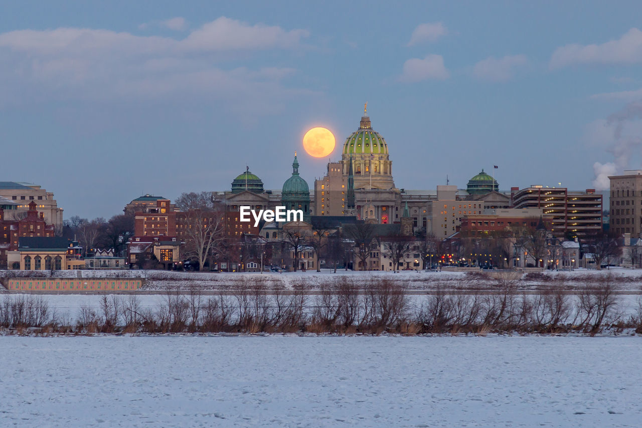 Illuminated buildings in city against moon during winter