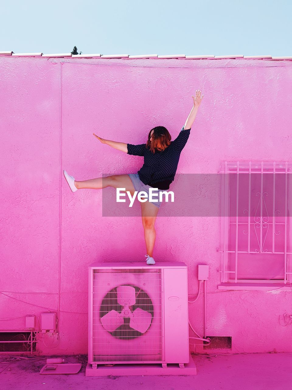 Woman exercising on air conditioner against pink wall during sunny day
