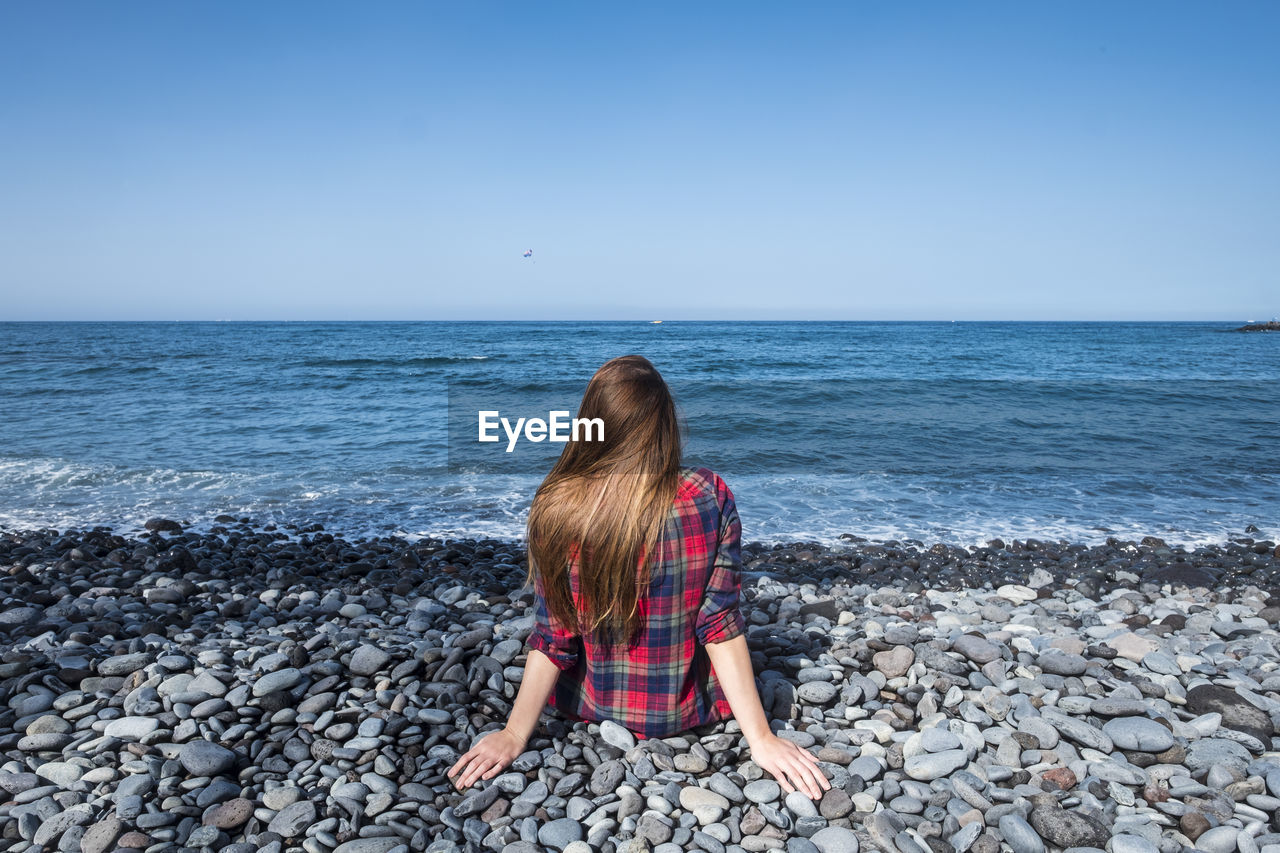 Rear view of woman sitting on rocks at beach against sky