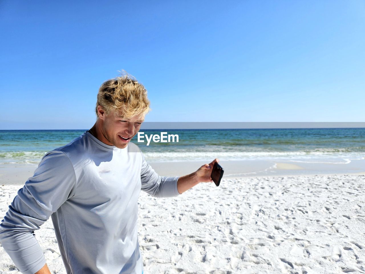 Young man millennial leaving sandy beach on vacation holding his cellphone.